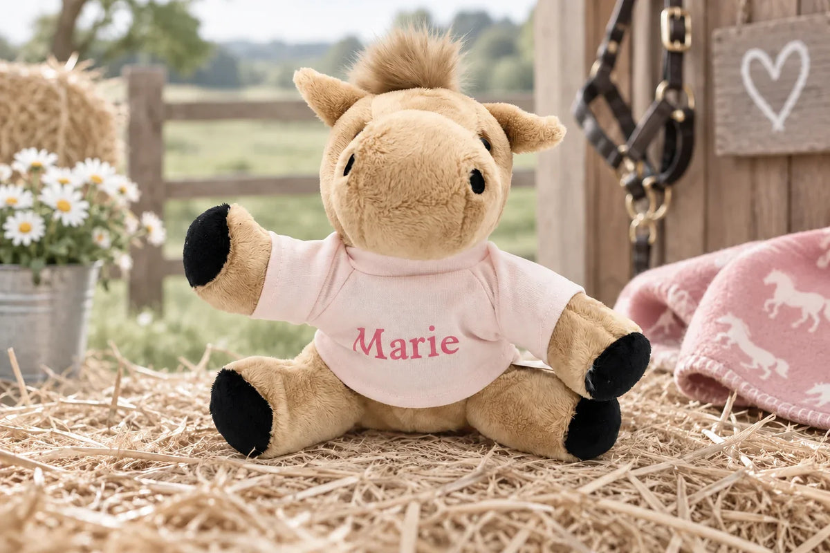 Beigefarbenes Kuschelpony mit rosa T-Shirt und dem Namen Marie auf Stroh in einer hellen Stallumgebung.